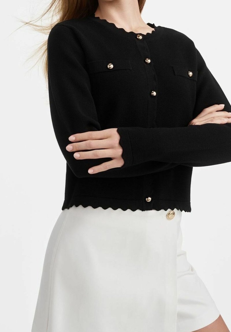 Femme portant un cardigan noir boutonné avec des bords festonnés et une jupe blanche avec un bouton doré, bras croisés sur la poitrine.