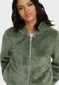 Zielona, futrzana bluza na zamek z kapturem, o miękkiej fakturze i srebrnym zamku błyskawicznym, zestawiona z białą bluzką pod spodem.