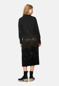 Schwarzer Pullover mit langen Ärmeln, kombiniert mit einem plissierten, knielangen Rock mit einem strukturierten Muster in Schwarz- und Bronzetönen. Stiefel mit dicker Sohle in Schwarz.