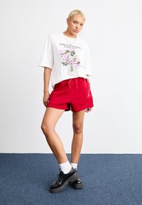 Bela oversized majica s cvetnim printom in napisom "JORDAN BRAND FOREVER", v kombinaciji z rdečimi kratkimi hlačami. Črne platforme in bele nogavice.
