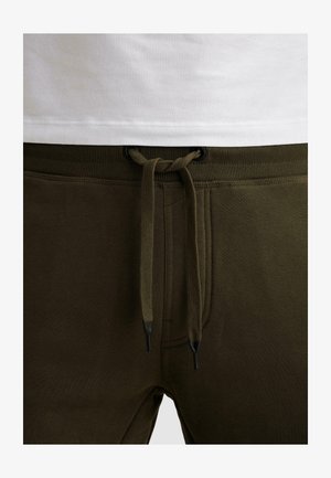 Zbliżenie na oliwkowozielone spodnie dresowe ze sznurkiem związanym kokardą w pasie, noszone z białą koszulą.