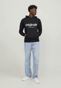 Joven de pie con las manos en los bolsillos, vistiendo una sudadera negra "Originals", jeans azul claro y zapatillas blancas, mirando hacia un lado.