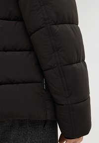 Manica di giacca a vento nera con sezioni trapuntate, tessuto liscio e sottile etichetta del logo vicino alla cucitura. Polsino testurizzato all'estremità.