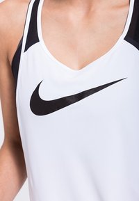 Débardeur Nike en blanc et noir. Il présente un décolleté en V profond, un tissu doux et un logo Swoosh noir bien visible au centre.
