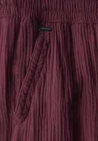 Bordeaux gekleurde stofbroek met een gekreukt patroon, voorzien van een elastische tailleband en een zijzak. Klein zwart merketiket zichtbaar.
