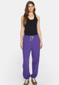 Kobieta stoi w czarnej bezrękawnik, fioletowych spodniach dresowych ze sznurkiem i brązowych trampkach na tle białego tła.
