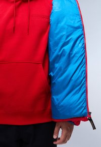 Sudadera roja con una manga acolchada azul, que cuenta con cierre de cremallera y un bolsillo frontal. La tela es suave, mientras que la manga tiene una textura brillante.