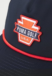 Casquette marine ornée d'un logo brodé orange et blanc affichant "PUMA GOLF U.S.A." Une corde rouge souligne le bord.