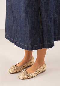 Bailarinas doradas de tejido con un detalle de lazo, tacón de madera bajo y un diseño abierto y texturizado, combinadas con una falda de mezclilla de un azul oscuro.