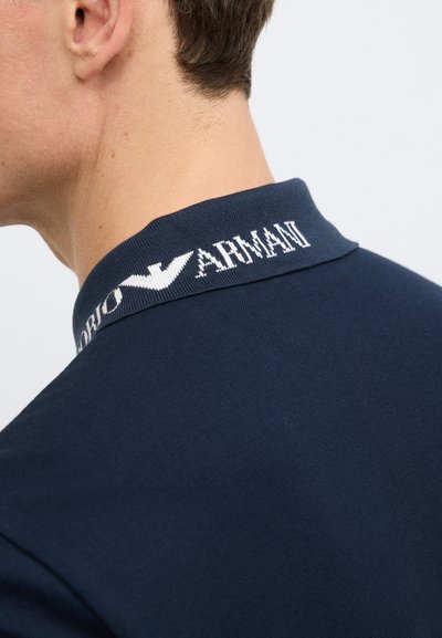 Tamsiai mėlynas polo marškinėlis su stačiakampiu klostuotu baltos spalvos „Armani“ logotipo siuvinėjimu ant apykaklės. Lygios tekstūros, klasikinio kirpimo, pabrėžiantis paprastą dizainą.