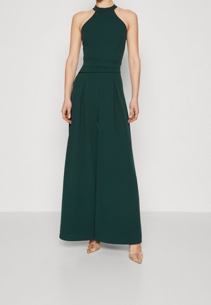 Vrouw die een mouwloze, donkergroene halter jumpsuit draagt met wijde pijpen en puntige, transparante hoge hakken met versieringen.