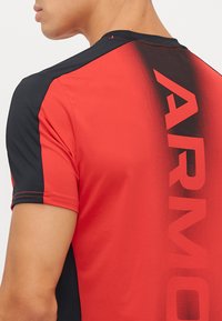 Rød og svart treningstrøye med gradientdesign. Har ordet "ARMOUR" i fet skrift langs ryggen. Korte ermer, glatt tekstur.