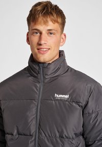 Giubbotto imbottito grigio con chiusura a zip, colletto alto e design a righe orizzontali. Presenta il logo "hummel" in bianco sul lato sinistro del petto.