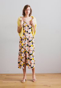 Vestido midi floral en rosa, amarillo y azul con un dobladillo con abertura; combinado con un suéter amarillo claro. Lleva sandalias de bloque con tacón en tonos neutros.