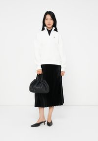 Donna che indossa un maglione bianco con zip, una gonna midi plissettata nera, décolleté neri, e tiene una borsa nera arricciata, in piedi davanti a uno sfondo bianco.