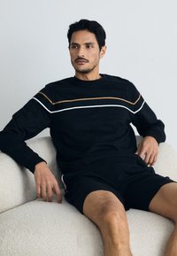 Fekete, hosszú ujjú póló, vízszintes barna és fehér csíkokkal, fekete sorttal párosítva. A modell egy textúrált, világos színű kanapén ül.