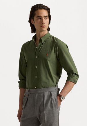 Chemise vert à col pointu avec un petit logo orange. Portée avec un pantalon gris ajusté et une montre marron.