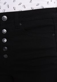 Czarne spodnie dżinsowe z wysokim stanem, zapinane na rząd pięciu metalowych zatrzasków. Tkanina ma gładką fakturę i minimalną rozciągliwość.