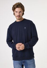 Sudadera azul marino en corte relajado, hecha de algodón suave, con un pequeño parche de logo en el pecho y puños y dobladillo acanalados.