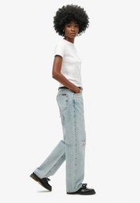 Jean déchiré bleu clair avec une coupe droite, associé à un t-shirt blanc ajusté et des chaussures noires. Modèle se tenant de côté.