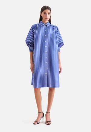 Kobieta ubrana w niebiesko-białą pionowo paskowaną sukienkę zapinaną na guziki z bufkami na rękawach i czarne sandały na wysokim obcasie, stojąca na białym tle.
