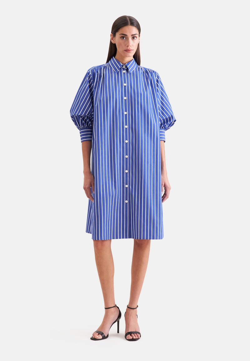 Femme portant une robe chemise à rayures verticales bleu et blanc avec des manches bouffantes et des sandales noires à talons hauts, debout sur fond blanc.