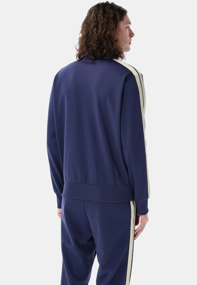 Giacca da tracksuit navy con strisce laterali chiare, polsini e orlo a costine. Tessuto liscio, taglio sportivo e colletto alto.