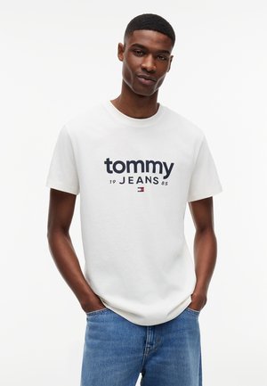 Muž v bielom tričku Tommy Jeans a modrých džínsach, stojaci s rukami v vreckách na jednofarebnom bielom pozadí.
