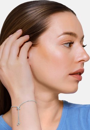 Młoda kobieta o jasnej karnacji i brązowych włosach, nosząca srebrną regulowaną bransoletkę, dotykająca swoich włosów, ubrana w niebieską koszulę, zwrócona w prawo.