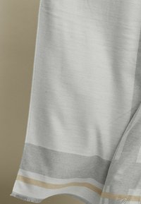 Světlý šedý textil s jemnou texturou, s vzorovaným lemem v šedých a zlatých pruzích. Okraje mají jemné třásně.
