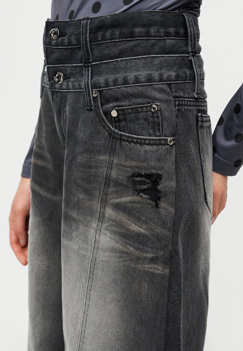 Jean en denim gris foncé taille haute avec un aspect d élavé et des détails usés. Comprend des fermetures à bouton et à glissière, plusieurs poches.