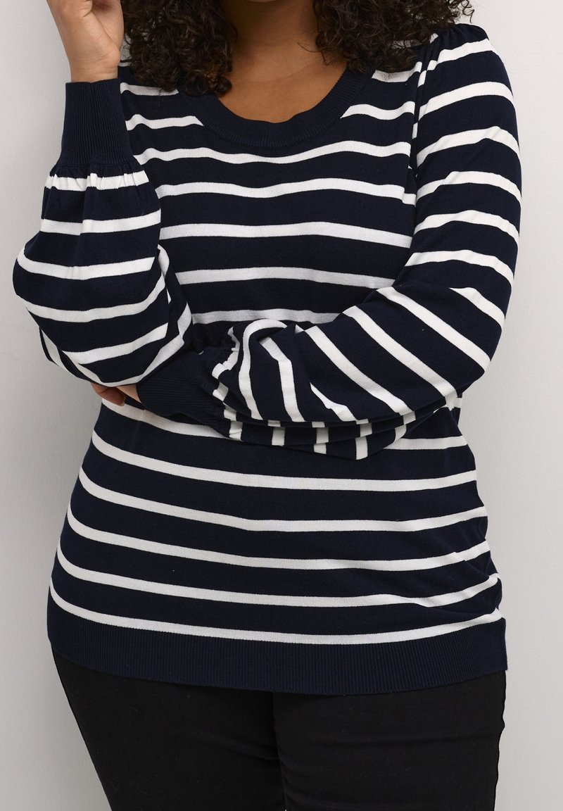 Femme portant un pull à manches longues à rayures horizontales bleu marine et blanches avec poignets et ourlet côtelés, assorti à un pantalon noir.