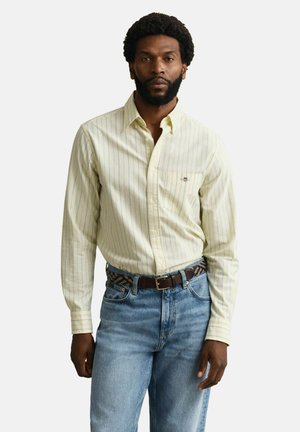 Homme barbu portant une chemise à boutons rayée jaune clair rentrée dans un jean bleu avec une ceinture à motifs, sur fond blanc.