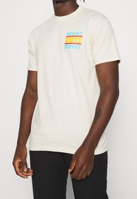 Krémové tričko, krátké rukávy, volný střih. Na levé části hrudi je grafika v modré, žluté a červené barvě: "REVOLT VOTE REPEAT".