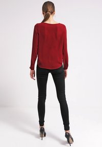 Femme avec une queue de cheval brune portant un chemisier rouge à manches longues, un jean skinny noir et des chaussures à talons hauts noires, debout de dos.