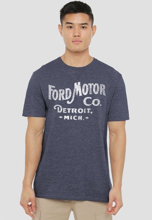 Young man wearing a navy blue Ford Motor Co. Detroit, Mich. graphic t-shirt and beige pants, standing against a gray background.