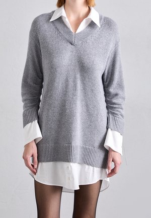 Femme portant un pull gris clair à col en V superposé sur une robe-chemise blanche boutonnée et des collants noirs, sur un fond uni.