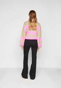 Stickad tröja med kraftiga rosa ränder på beige bakgrund, kombinerad med svarta, figursydda utsvängda byxor, sedd bakifrån.
