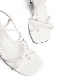 Weiße Riemchensandalen mit quadrater Zehenpartie und dünnen Lederbanden, die über den Fuß verlaufen, auf einem weißen Hintergrund präsentiert.