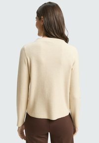 Jasnobeżowy sweter z okrągłym dekoltem, długimi rękawami i lekko zaokrąglonym dołem, wykonany z miękkiego materiału dzianinowego. Teksturowane wykończenie.