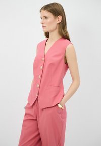 Gilet sans manches rose avec un décolleté en V profond, cinq grands boutons dorés et deux poches latérales, associé à un pantalon taille haute assorti.