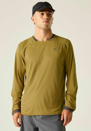 Mann mit olivgrünem Langarmshirt mit Netz-Schulterpartien und schwarzer Kappe, steht vor einem schlichten hellen Hintergrund.