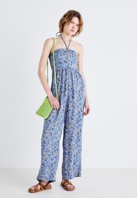 Modra cvetna kombinezonska obleka brez naramnic, z nabranim pasom in širokimi hlačami. V kombinaciji z zeleno tkano torbico in rjavimi sandali.