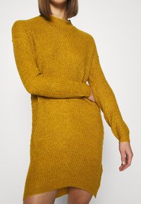 Abito maglione in giallo senape, caratterizzato da un colletto alto, maniche lunghe e una vestibilità rilassata con texture a coste e spacco laterale.