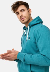 Sweat à capuche zipé teal avec une grande capuche et des cordons blancs, présentant un logo sur la poitrine. Le tissu semble doux et texturé.
