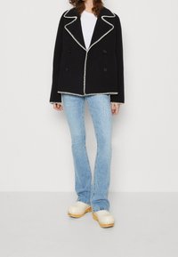 Musta kaksirivinen takki, jossa on erottuva valkoinen tikkaus, leveät kaulukset ja levenevät hihat, yhdistettynä vaalensinihin farkkuihin ja beigeihin puutöppösiin.