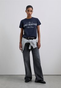 Camiseta azul marino con la inscripción "SPORT HERITAGE" en blanco, combinada con jeans de pierna ancha en gris. Una sudadera gris claro está anudada alrededor de la cintura.