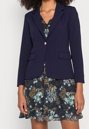 Femme portant un blazer bleu marine foncé avec un seul bouton sur une courte robe fleurie avec des motifs de feuilles bleues, vertes et beiges.