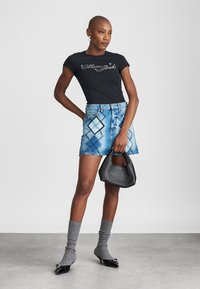 T-shirt noir ajusté avec le logo "Marc Jacobs", mini-jupe en denim bleu avec un motif en diamant, chaussettes en tricot grises et un petit sac à main noir.