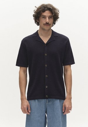 Mann mit lockigem Haar und Schnurrbart, trägt ein schwarzes kurzärmeliges Hemd mit Knöpfen und hellblaue Jeans, steht vor einem einfarbigen weißen Hintergrund.